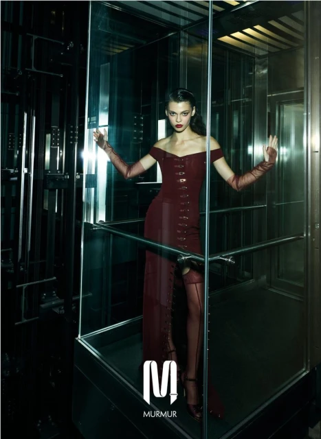 Fashion shot of a woman in a burgundy dress posing inside a mirrored elevator with long gloves, stockings, and heels, set against a dark interior.