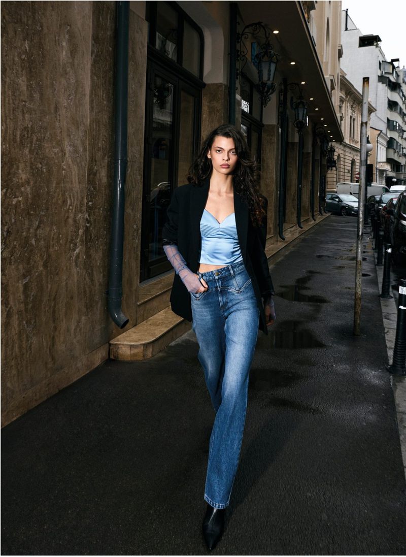 A fashionable woman in a blue top, blazer, and jeans walks along a city sidewalk, showcasing a blend of casual and chic style.