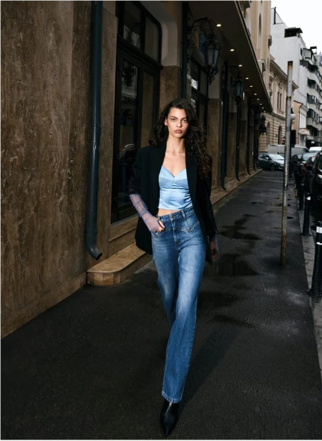 A fashionable woman in a blue top, blazer, and jeans walks along a city sidewalk, showcasing a blend of casual and chic style.