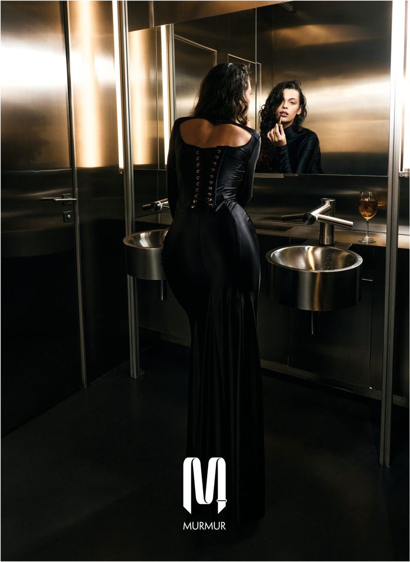 An adult woman in a long black dress stands in a modern bathroom looking in the mirror while applying lipstick, captured in a medium shot.