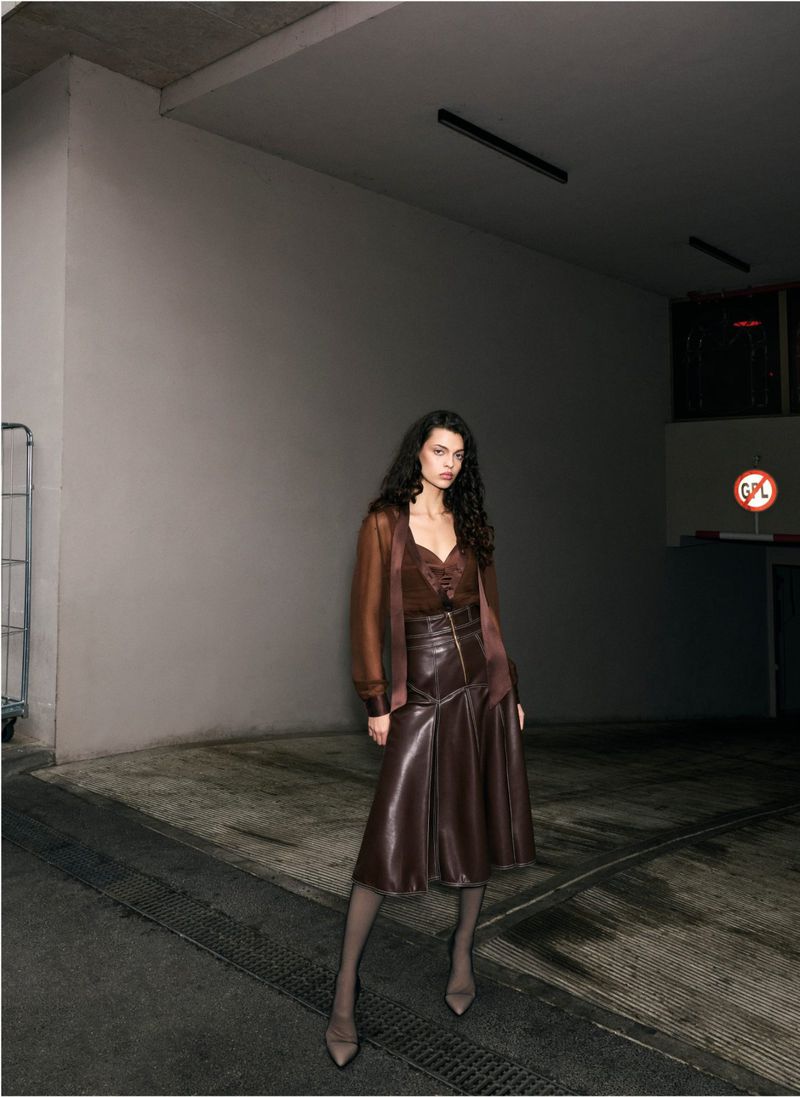 Adult female posing indoors against a neutral background, wearing a brown leather skirt and top with long sleeves, accessorized with heels.
