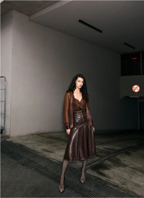 Adult female posing indoors against a neutral background, wearing a brown leather skirt and top with long sleeves, accessorized with heels.