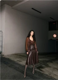 Model posing in a brown sheer blouse and chocolate brown leather midi skirt inside a concrete underground parking garage.