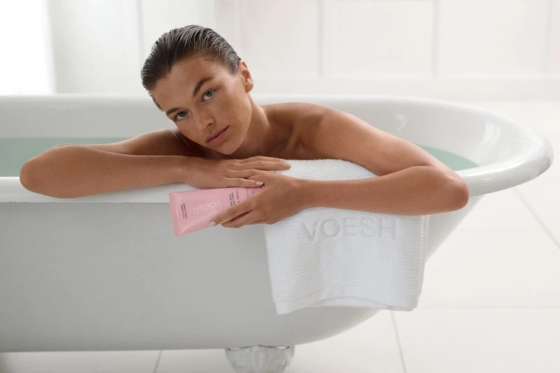 Woman relaxes in a white bathtub, holding a pink VOESH beauty product, with a matching white VOESH towel in a light-filled bathroom.