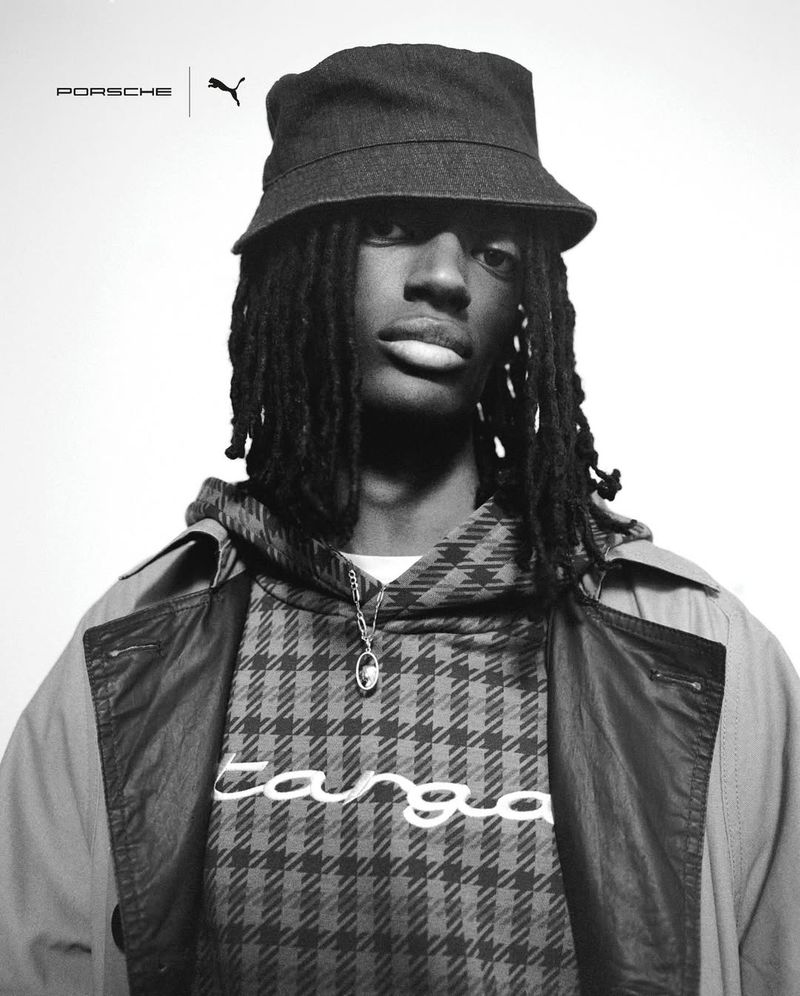 Black and white portrait of Jahmal John Baptiste in a bucket hat and houndstooth hoodie against a light studio backdrop.