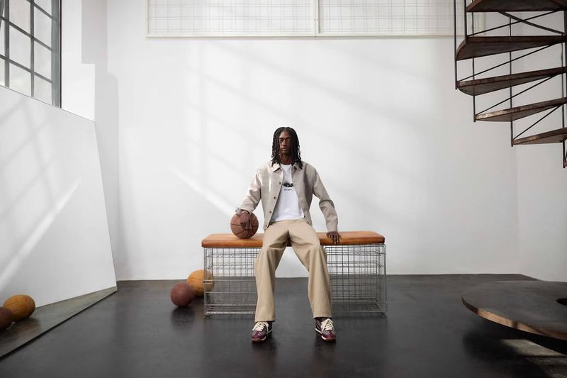 An adult male sits on a bench in a photo studio holding a basketball, wearing a jacket, pants, and shoes; there are several basketballs nearby.