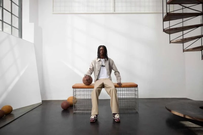 An adult male sits on a bench in a photo studio holding a basketball, wearing a jacket, pants, and shoes; there are several basketballs nearby.