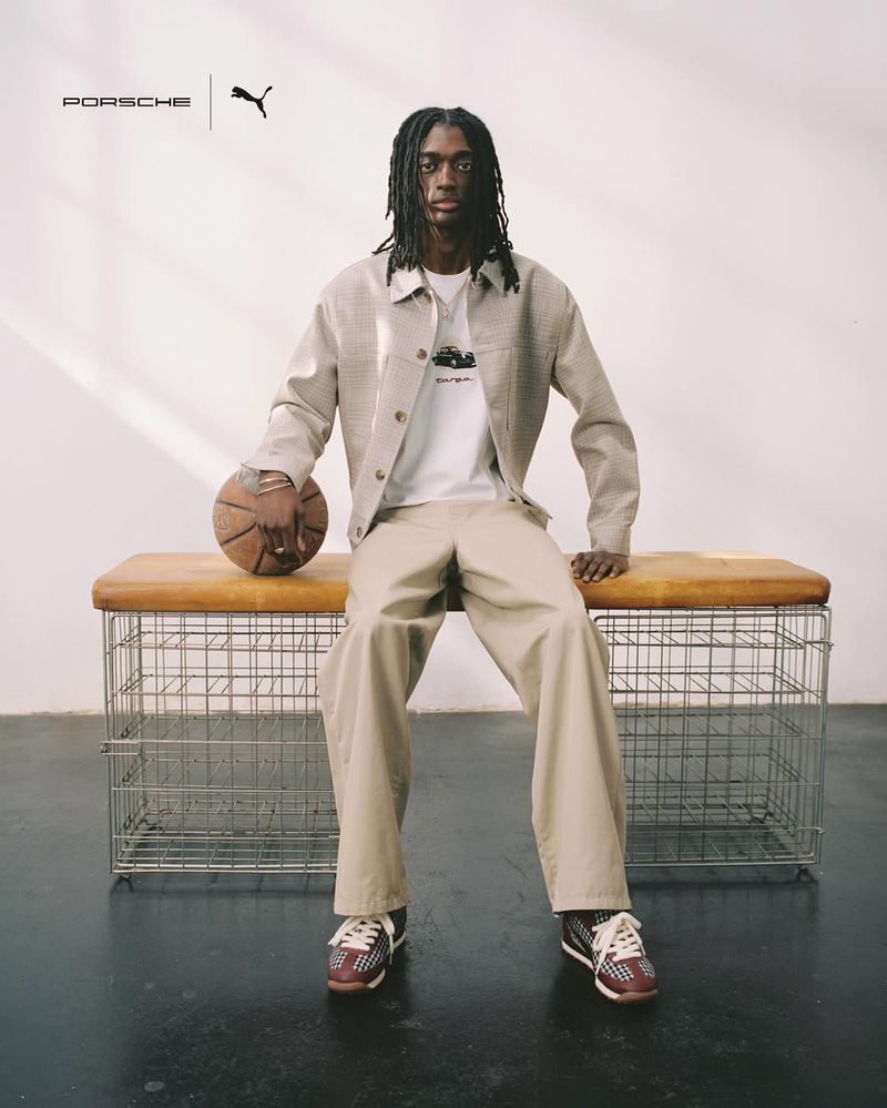 An adult male sits on a bench indoors wearing neutral-colored clothing and athletic shoes, holding a basketball, with logos in background.
