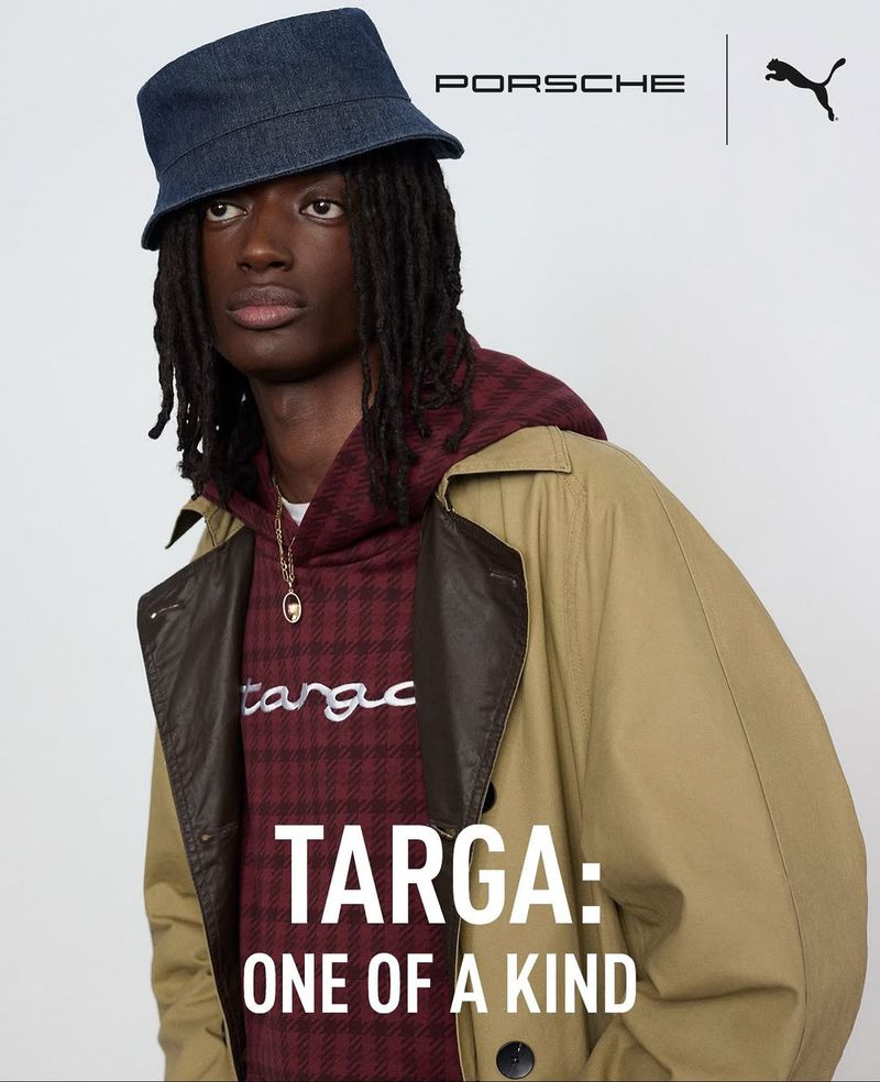 Portrait of a Black male model in a denim bucket hat, burgundy plaid hoodie, and tan trench coat, showcasing fashion apparel in a studio.