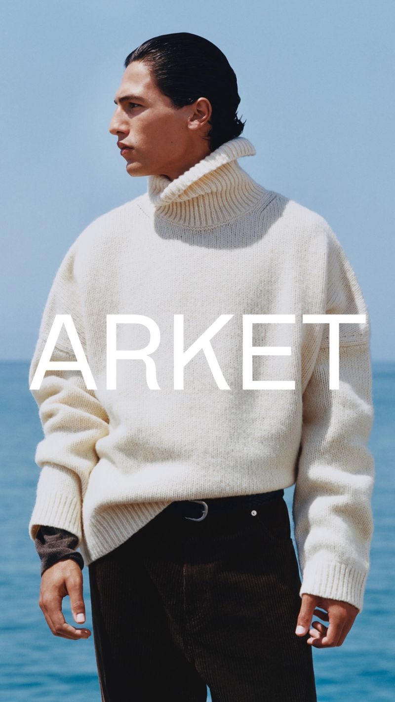 A man in a cream turtleneck sweater poses in front of the ocean, showcasing a fashion-forward aesthetic with natural lighting and minimalist style.