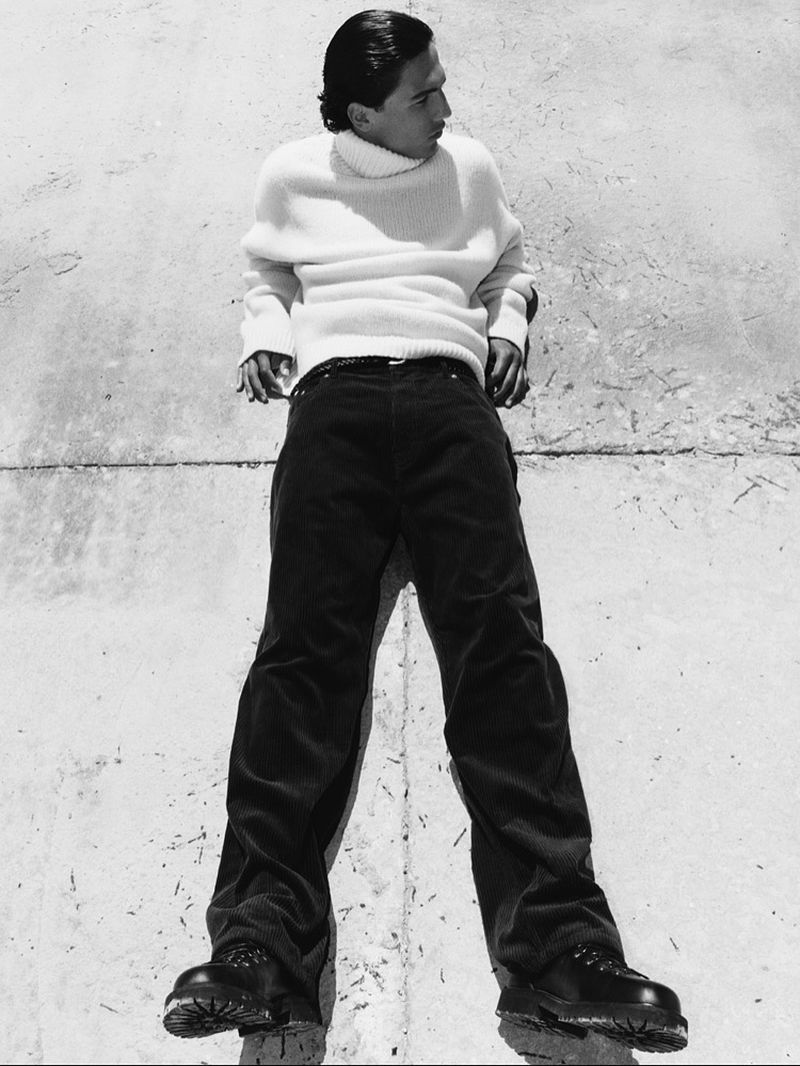 Full-length portrait of a young adult male in a white turtleneck sweater, dark trousers, and black boots, lying on a concrete surface.