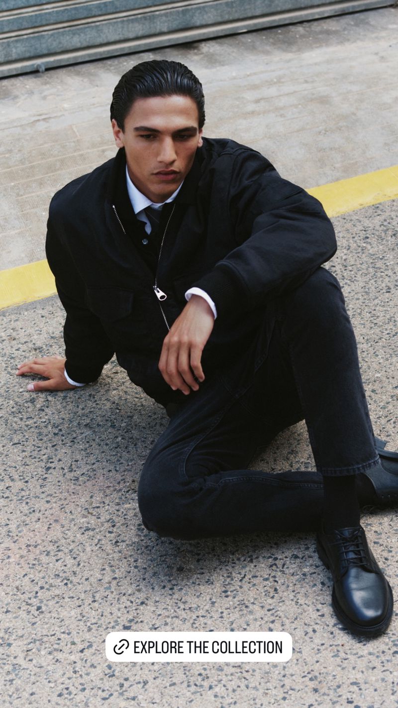 Male model in a black jacket and jeans sitting on a concrete surface, part of a fashion collection, captured in natural lighting.