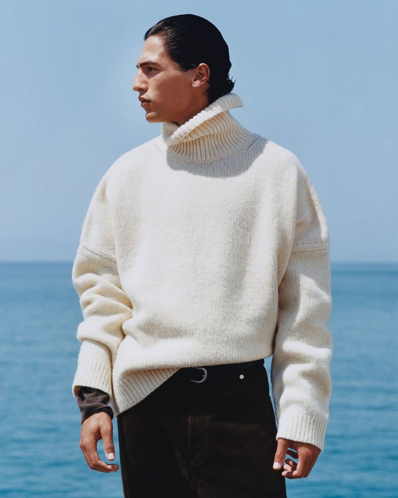 Man in a cream turtleneck sweater and dark pants stands outdoors with a blue sky and the sea in the background in a medium shot.