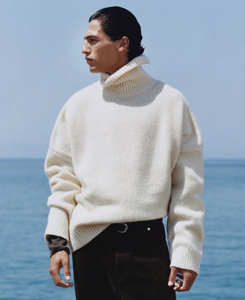 A fashionable adult male stands outdoors against the sea and sky, wearing a white turtleneck sweater, brown pants, and a black belt.