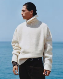 A male model wearing an oversized cream knit turtleneck sweater standing against an open ocean and clear sky background.