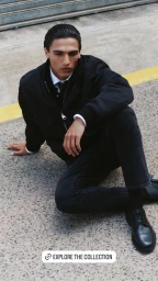 Model Morgan Sahadow sitting on concrete wearing a black zip jacket, white collared shirt, black trousers, and leather shoes.