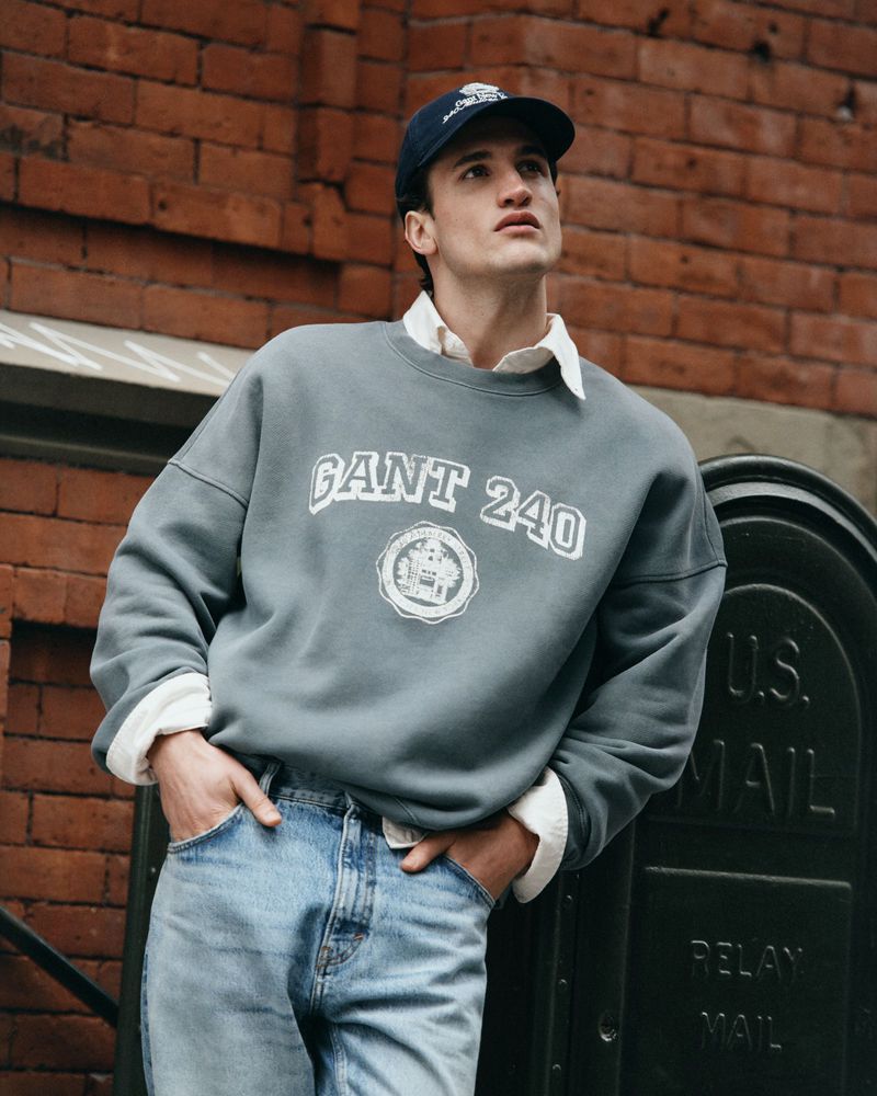 Young adult male in casual fashion poses against a brick wall wearing jeans, a gray 'GANT 240' sweatshirt and baseball cap.