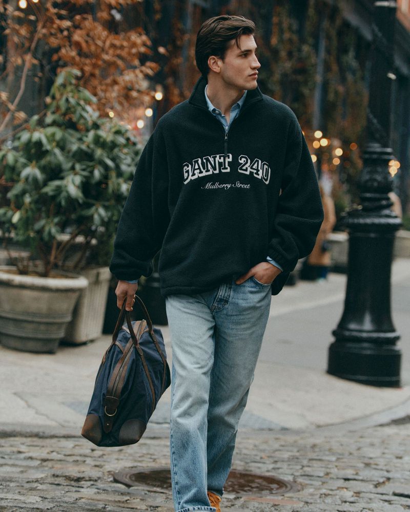 A young man wearing a GANT sweatshirt and jeans walks down a brick street carrying a blue duffel bag on a city street with trees.