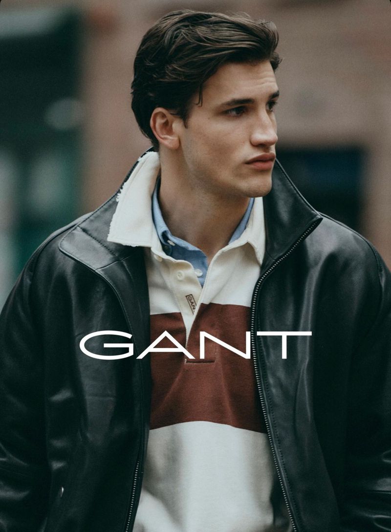 A young adult male poses in a black leather jacket and rugby shirt, showcasing a casual fashion aesthetic against an urban backdrop.