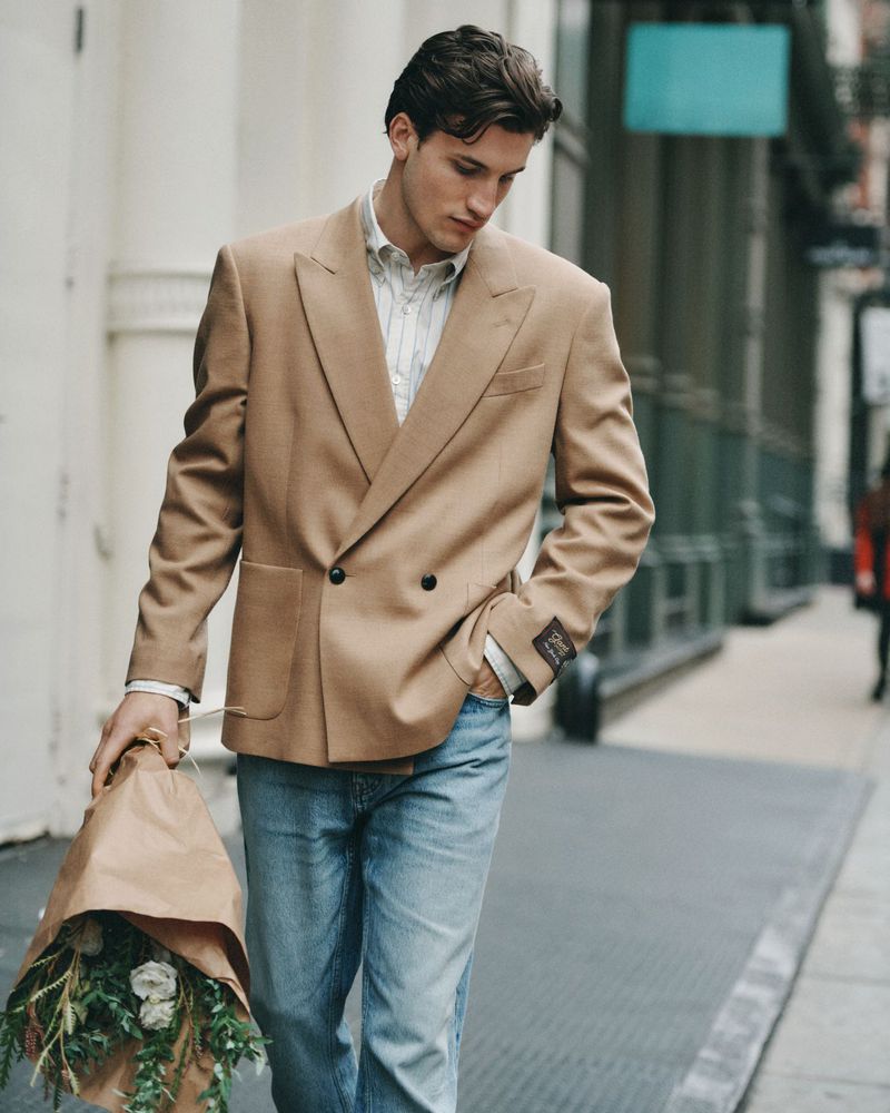 A young adult male in a tan blazer and jeans walks down a city sidewalk holding a bouquet of flowers in a brown paper bag.