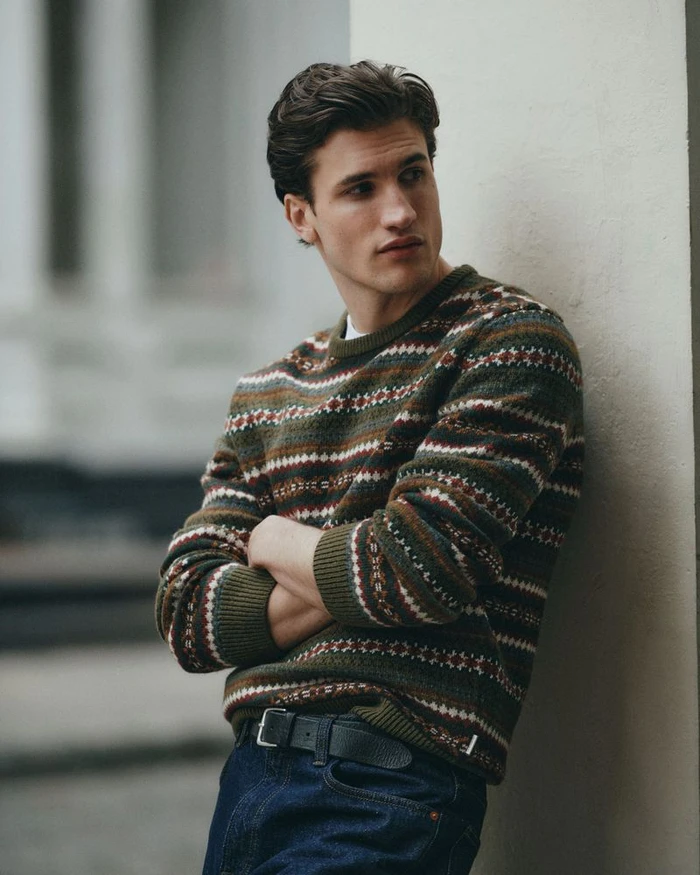A man wearing an olive patterned sweater and denim jeans leaning against a wall outdoors