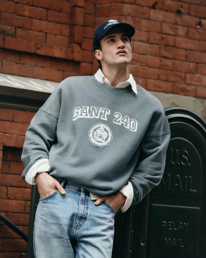 Man wearing a grey Gant collegiate sweatshirt, white collared shirt, denim jeans, and a navy baseball cap leaning against a brick wall.