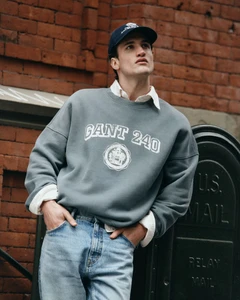 Man wearing a grey Gant collegiate sweatshirt, white collared shirt, denim jeans, and a navy baseball cap leaning against a brick wall.