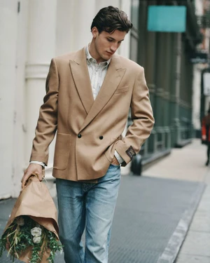 A man in a double-breasted camel blazer and blue jeans walks along a city street carrying a wrapped bouquet of flowers.