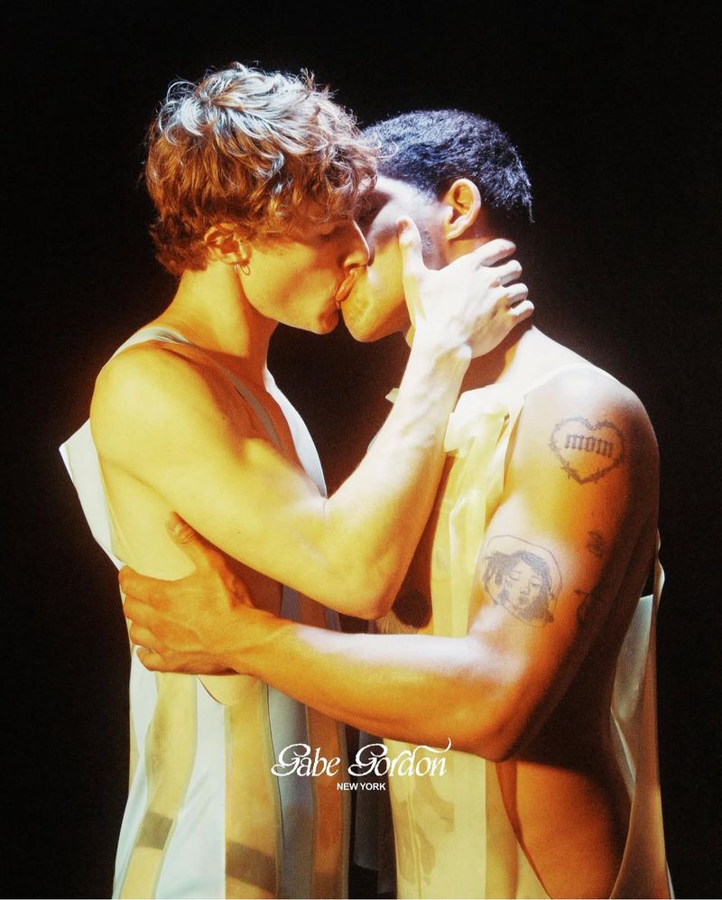 Two men are close together in a warmly lit studio, embracing and kissing, both wearing open white tops. The background is dark.