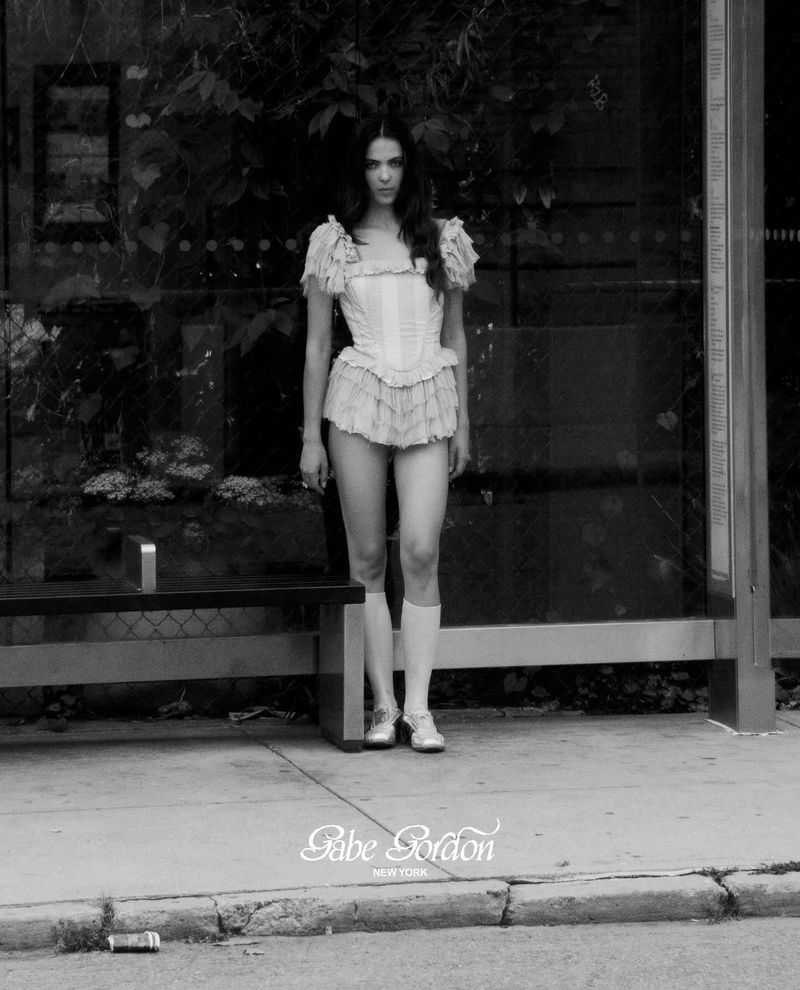 Model standing on a sidewalk near a bus shelter wearing a ruffled top and matching shorts in a black and white editorial campaign image.