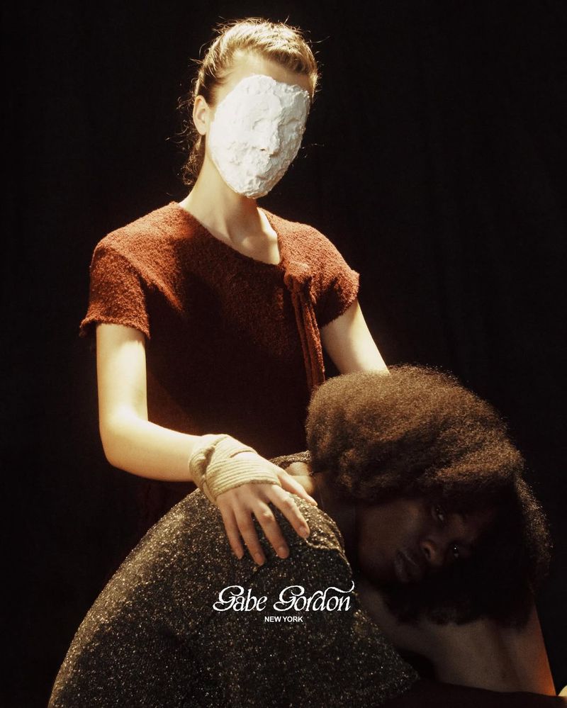Two women pose in dim light. One woman has a face mask and is touching the other woman's shoulder. Fashion editorial photography.