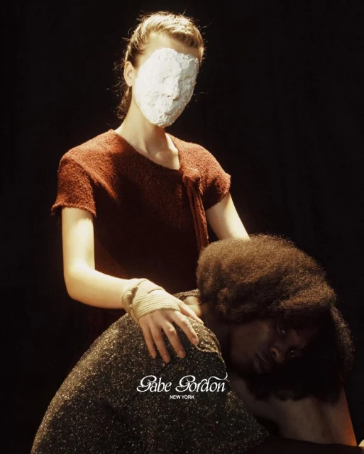 Two women pose in dim light. One woman has a face mask and is touching the other woman's shoulder. Fashion editorial photography.