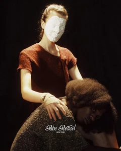 Model wearing a textured rust knit top and white plaster face mask placing a bandaged hand on another model with an afro.