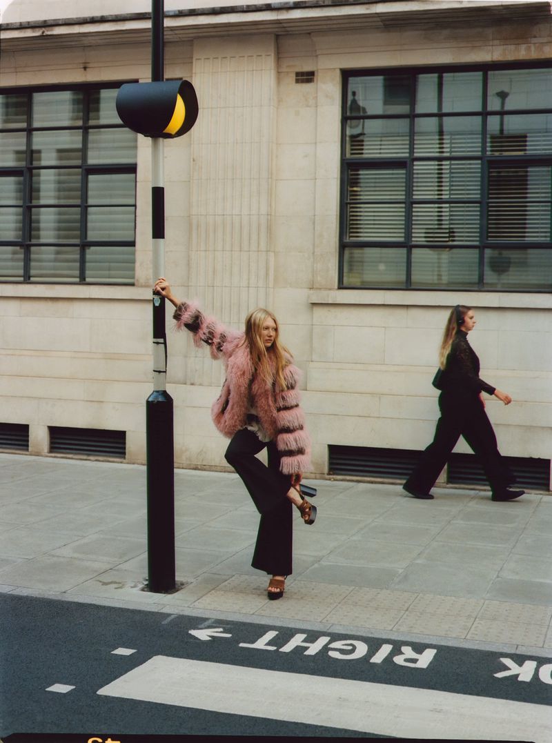 A woman in a pink coat poses near a traffic signal on a city street, while another woman walks in the background.