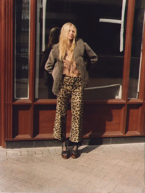 A fair-skinned woman with blonde hair stands outdoors in leopard-print pants, a gray faux fur coat, and platform heels in a fashion shot.