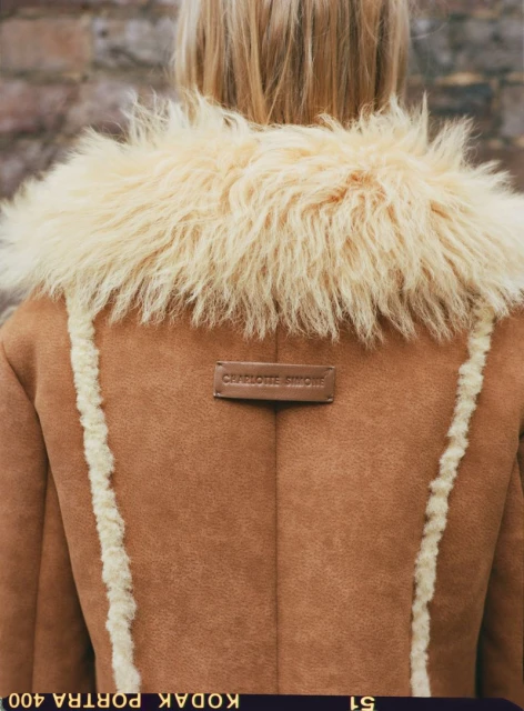 Blonde woman wearing a light brown coat with beige faux fur collar and trim, shot from the rear against a brick wall.