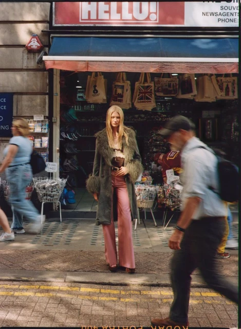 A fashion photo in an urban setting features a slender woman in a fur coat and pink pants standing outside a store with pedestrians.