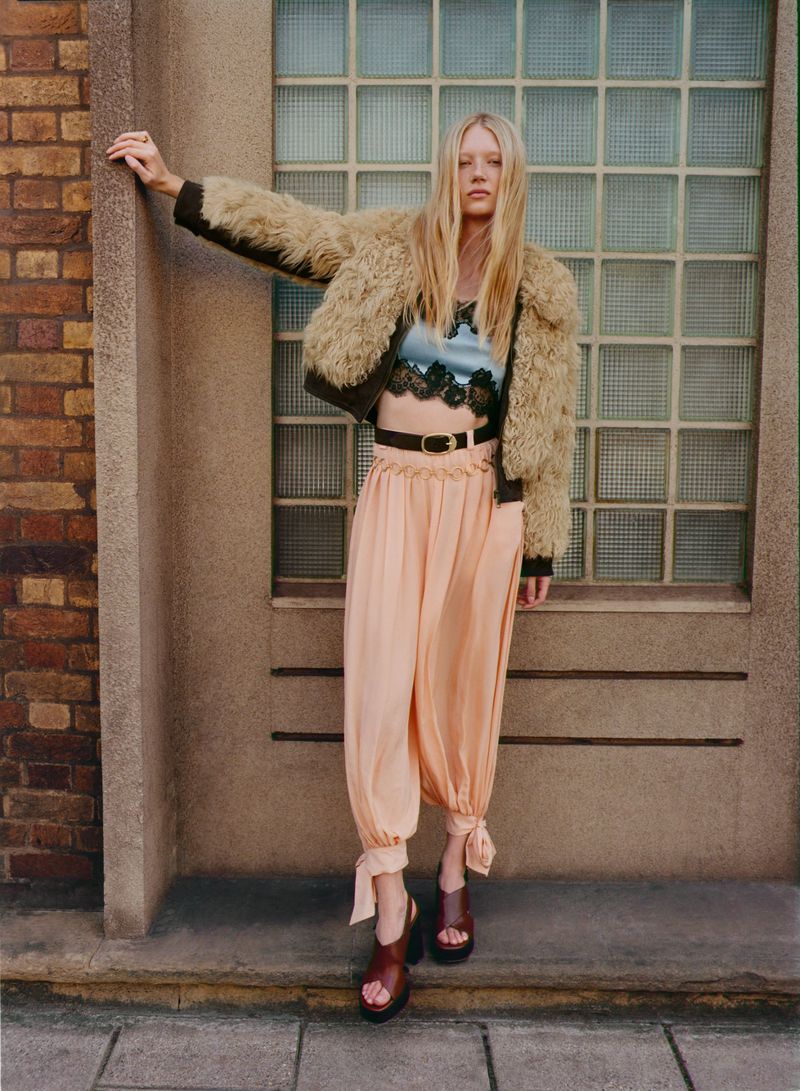 Fashion shoot of a woman wearing a fur jacket, blue crop top, pink pants, and heels, posing against a brick and concrete wall backdrop.