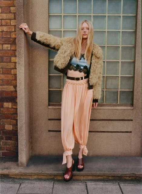 Fashion shoot of a woman wearing a fur jacket, blue crop top, pink pants, and heels, posing against a brick and concrete wall backdrop.