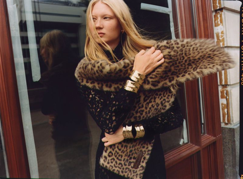 A blonde-haired woman in a leopard-print scarf and gold bracelets stands near a window, showcasing a dark, stylish fashion ensemble.