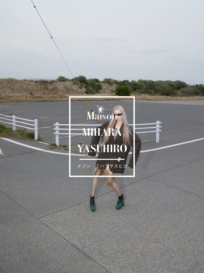 A fashion shot on an outdoor runway featuring a stylish woman in a blazer, fishnet stockings, and boots against a grey asphalt backdrop.