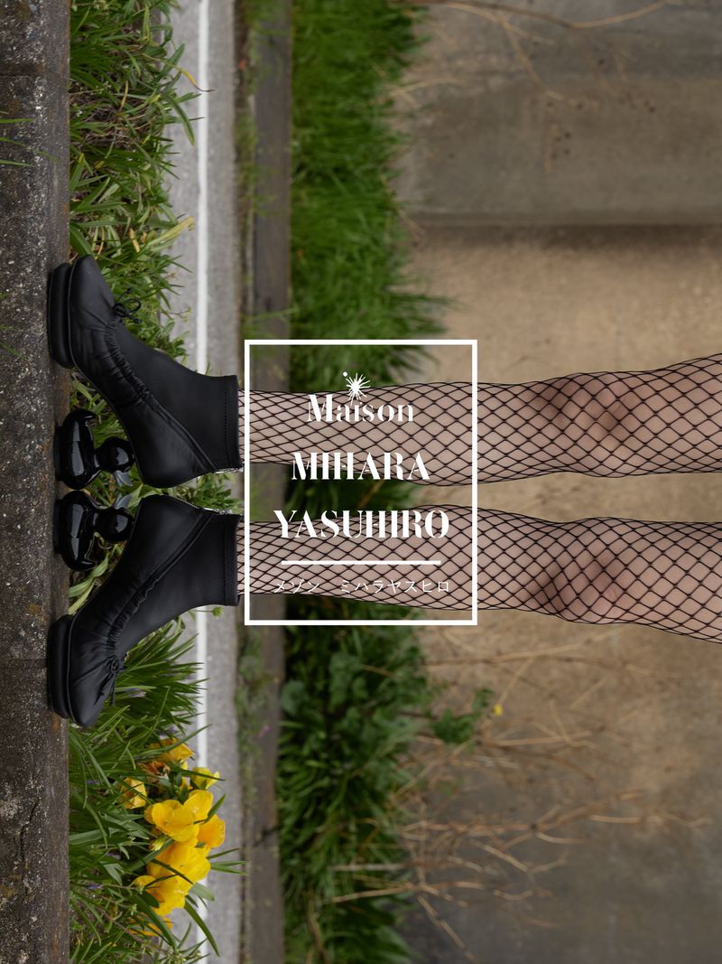 Close-up of a person wearing black platform boots and fishnet stockings on a sidewalk with grass and flowers, brand logo visible.