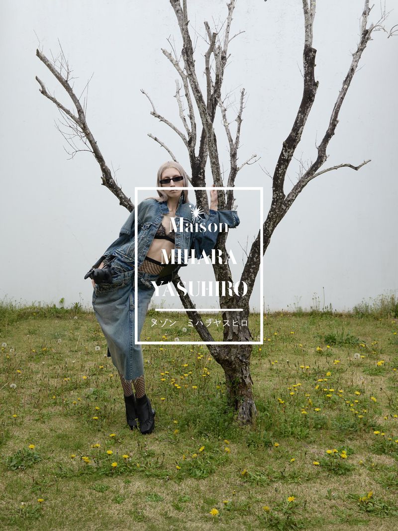 Fashionable blonde woman in jean jacket and skirt poses near tree in outdoor editorial shot. Designer brand Maison Mihara Yasuhiro is visible.