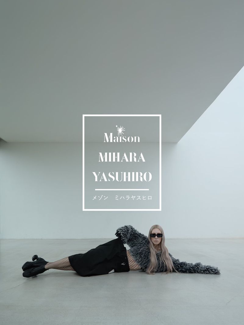 Fashion photography featuring a woman lying on the floor in a gray, minimal space, wearing a fur coat, sunglasses, and black shorts.