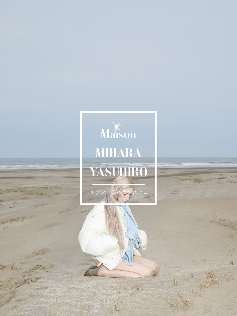 Blonde woman kneeling on a sandy beach wearing a white puffer jacket and blue scarf against an ocean backdrop.