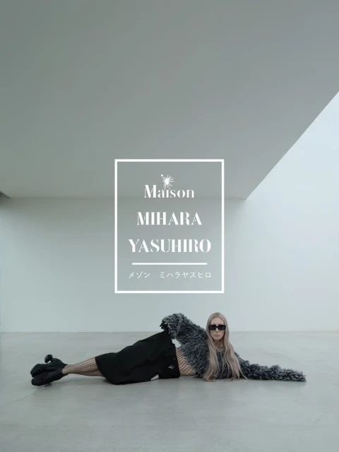 Model wearing dark sunglasses and a textured grey cropped sweater lying horizontally on a concrete floor in a minimalist studio.