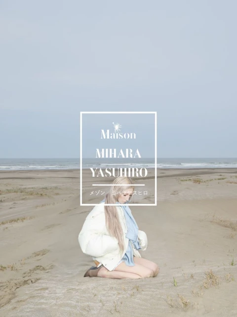 Blonde woman kneeling on a sandy beach wearing a white puffer jacket and blue scarf against an ocean backdrop.