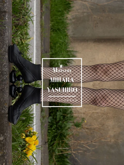 Model wearing black fishnet stockings and black boots resting feet near a curb with yellow flowers.