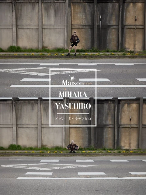 Model standing against a weathered concrete wall and asphalt road for the Maison Mihara Yasuhiro campaign.