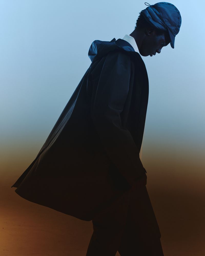Fashion shot of dark-skinned adult male wearing a hooded raincoat and baseball cap, looking down against a blue gradient background.